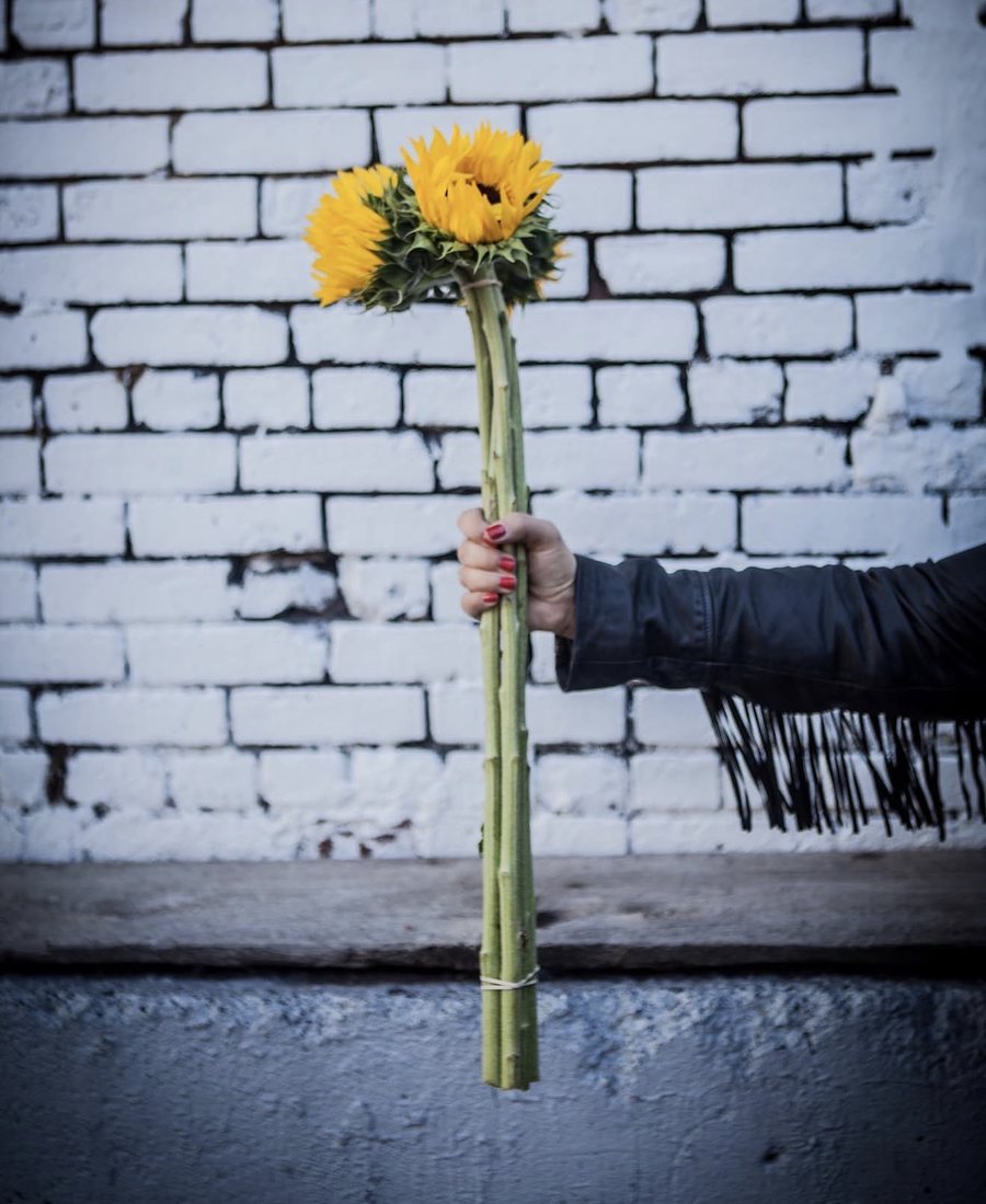 Jennifer’s hand, in a fringed leather jacket, holding out a bunch of sunflowers against a white brick wall