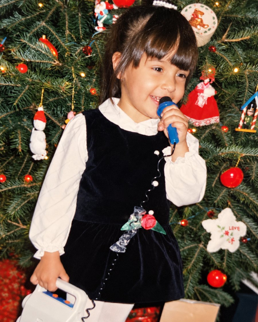 Jennifer as a little girl in a velvet holiday dress, singing into a toy microphone in front of a Christmas tree